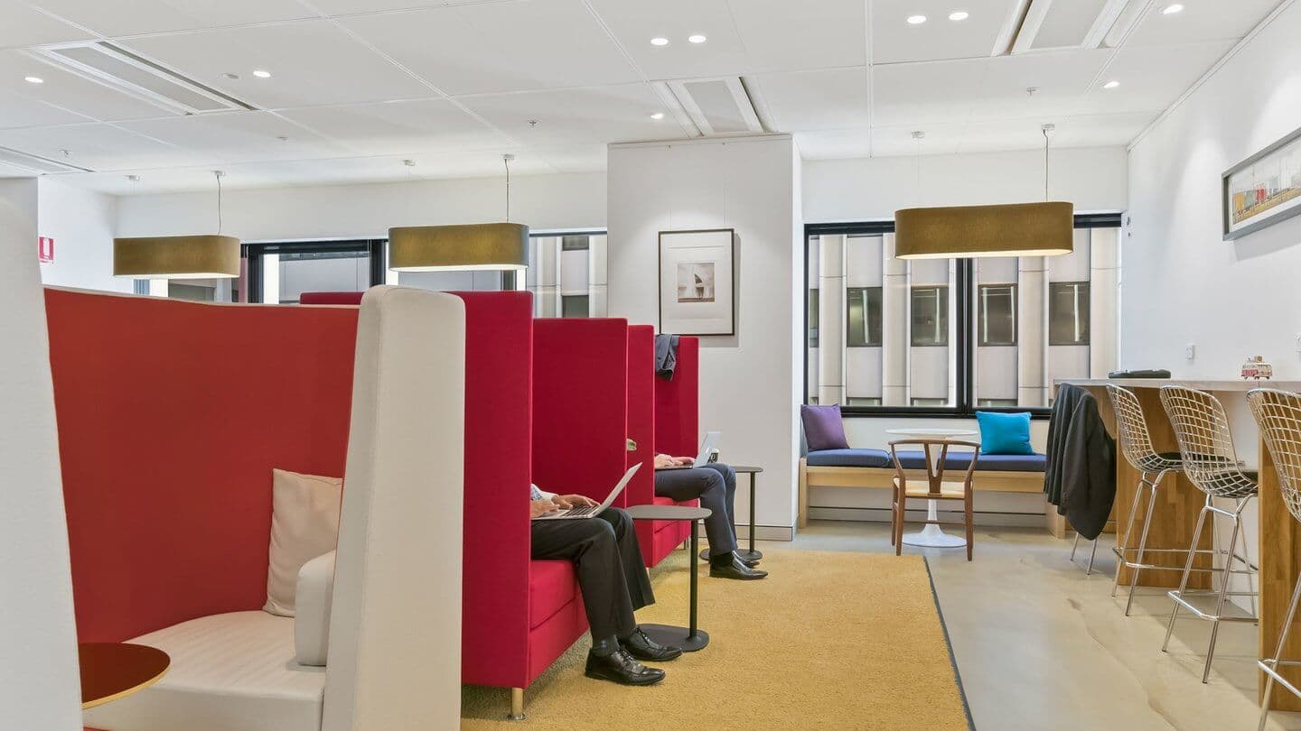 Modern office lounge with red cushioned booths and light wooden tables. A person sits in one booth with a laptop. Tall windows let in natural light. Bar stools and pendant lights add to the contemporary decor. Neutral and red tones throughout.