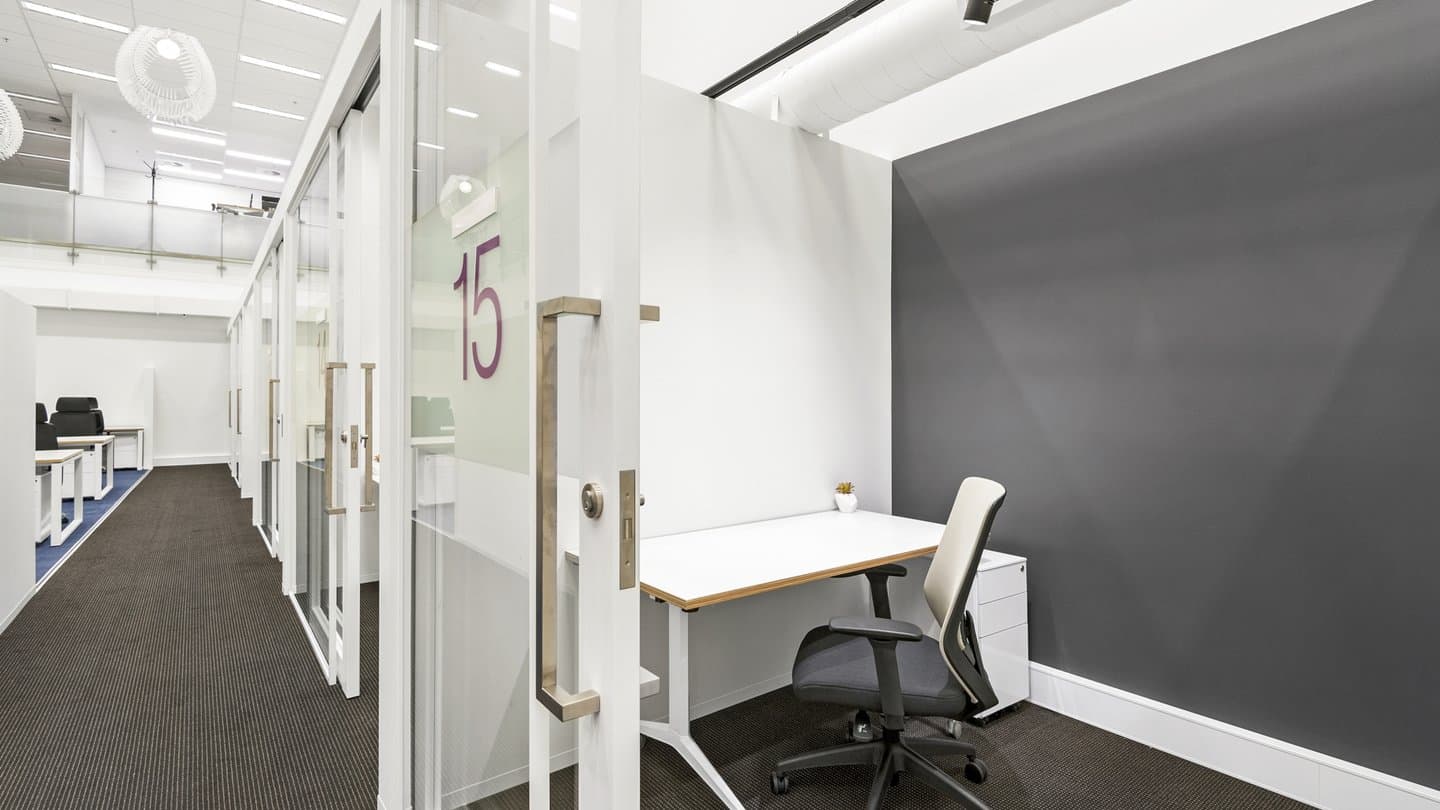 A modern office space with individual workstations featuring glass partitions and white desks. One desk has a white chair and small potted plant. The hallway has a dark carpet and leads to more workspaces in the background.
