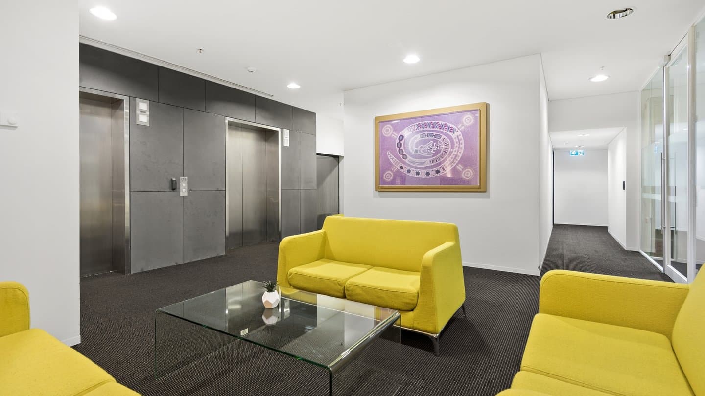 Lobby with yellow sofas and a glass coffee table. There are two elevators with metallic doors on a gray wall. A framed artwork with purple tones hangs on a white wall. The floor is carpeted in dark gray.