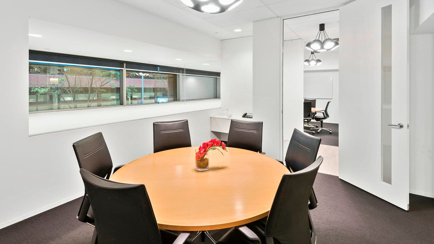 Modern office meeting room with a round wooden table surrounded by six black chairs. A small decorative plant is in the center. Large windows show an outside view, and a doorway on the right leads to another room with similar lighting.