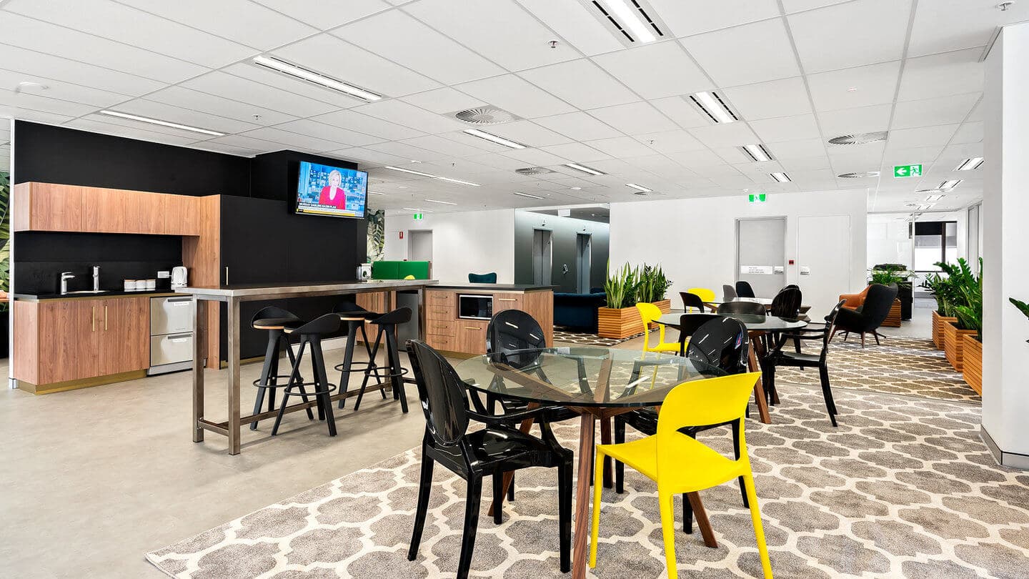 Modern office break room with glass tables, black and yellow chairs, a bar-height counter with stools, a kitchenette with wood cabinets, and a wall-mounted TV. Potted plants and patterned rugs decorate the space, creating a welcoming atmosphere.