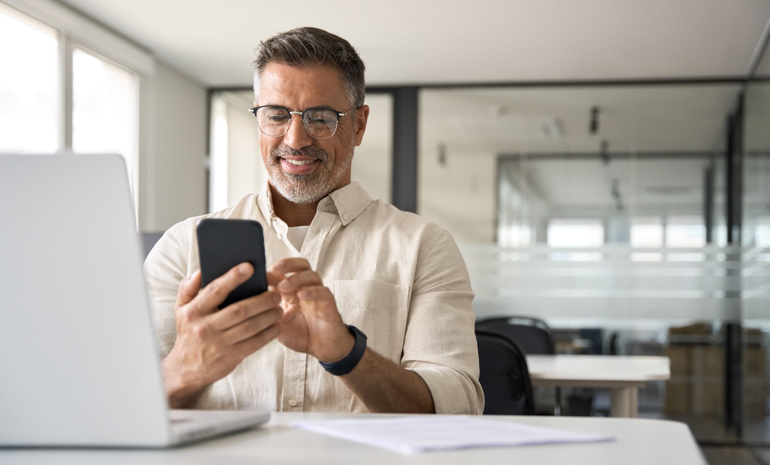 Middle aged Hispanic business manager ceo using cell phone mobile app, laptop. Smiling Latin or Indian mature man businessman holding smartphone sit in office working online on gadget with copy space.
