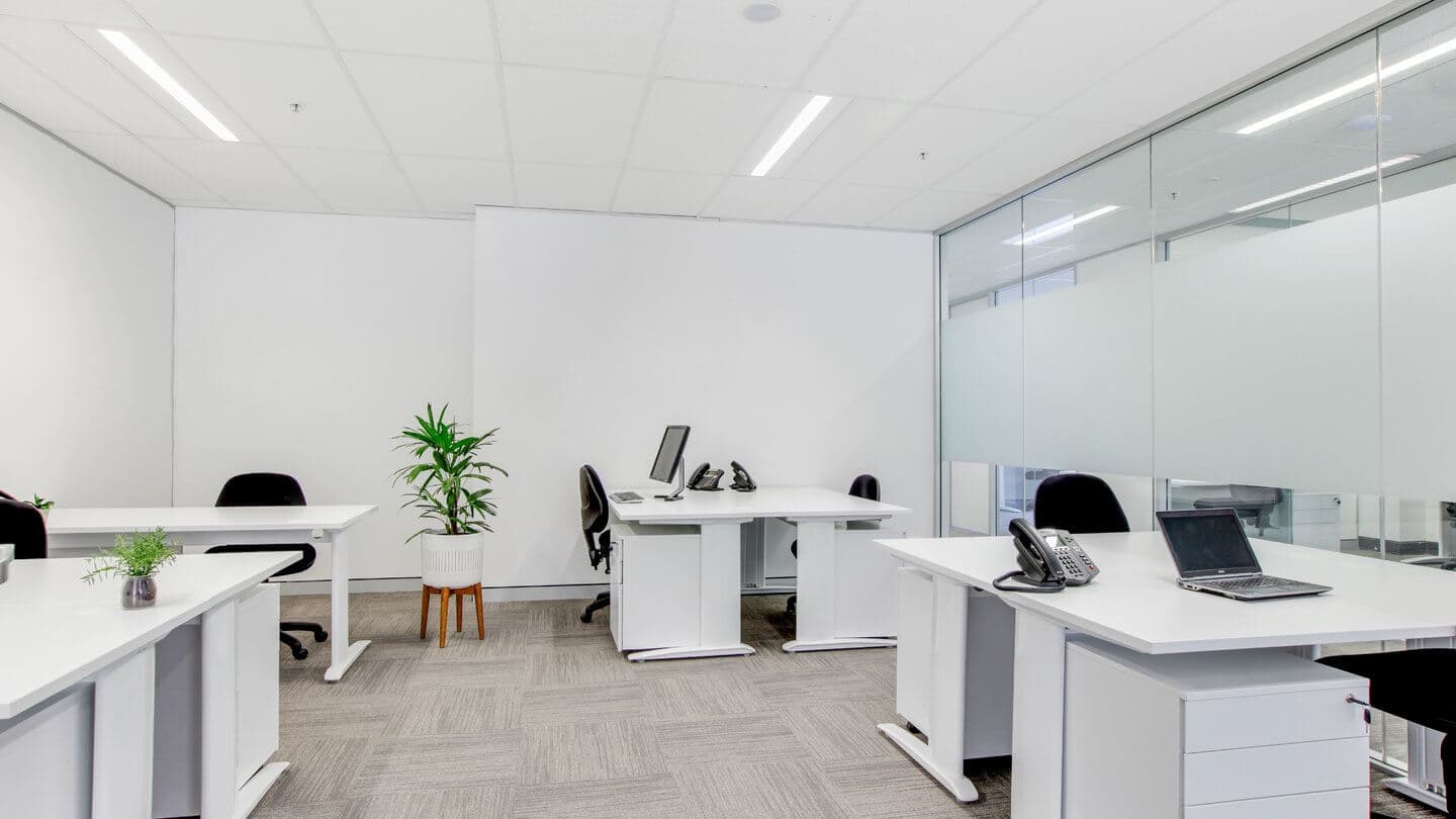 A modern office with white desks and black chairs. There are telephones, computers, and a laptop on the desks. A potted plant is on the floor, and a small plant is on one desk. The room features glass walls and a carpeted floor.