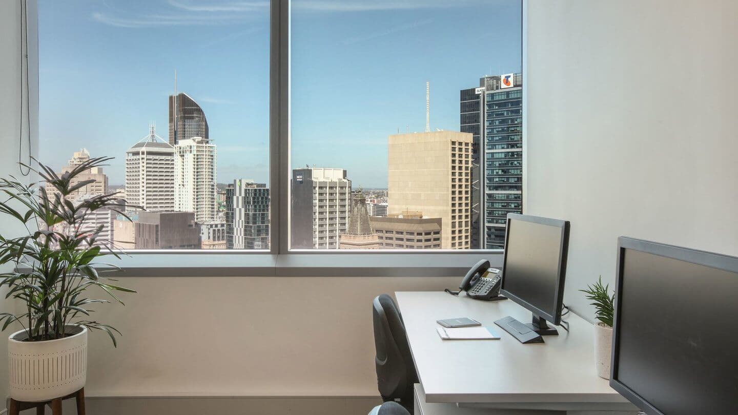 A modern office with two desks, each equipped with a computer monitor, phone, and notepads, near large windows offering a cityscape view. The room includes potted plants, and the skyline features several tall buildings under a clear blue sky.