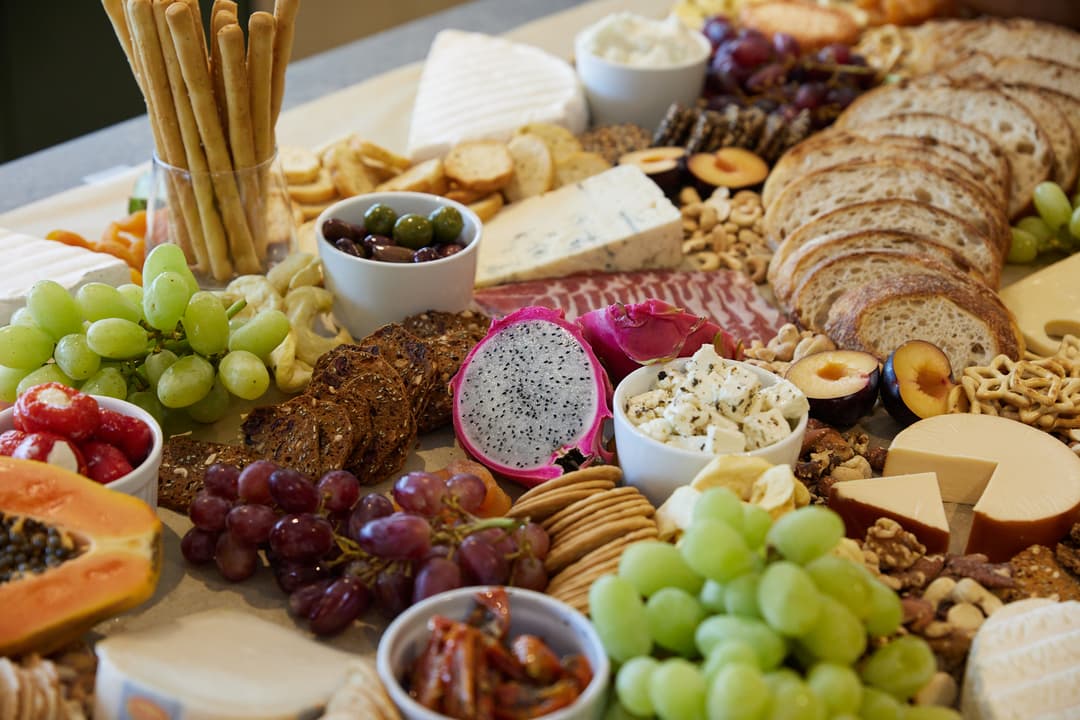 A charcuterie board filled with assorted cheeses, slices of bread, crackers, grapes, dragon fruit, olives, nuts, cured meats, breadsticks, and various dips. The arrangement is colorful and abundant.