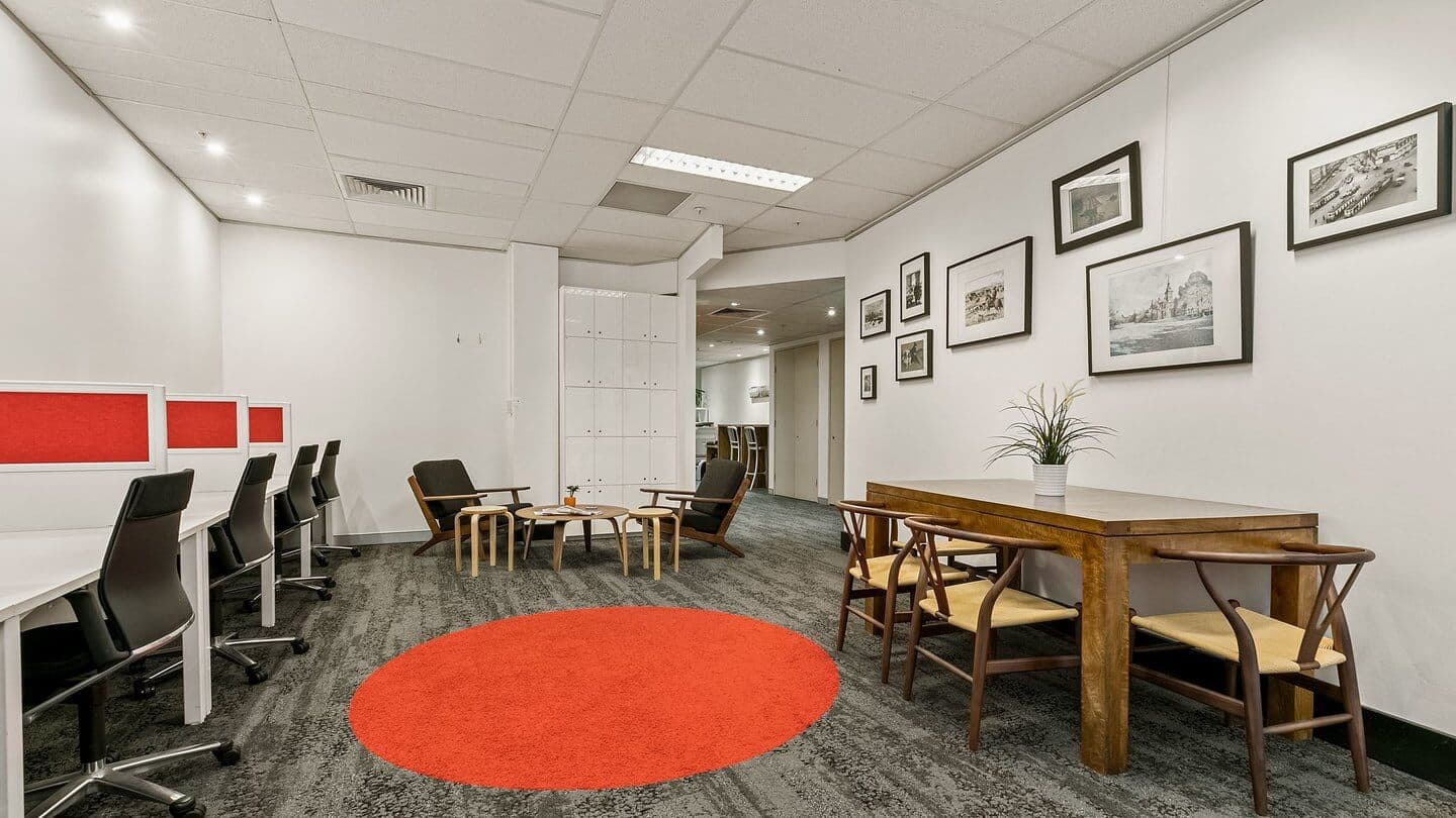 Modern office space featuring a row of desks with black chairs, a wooden table with four chairs, and a seating area with armchairs and a coffee table. The floor has a red circular rug, and black-and-white photos decorate the walls.