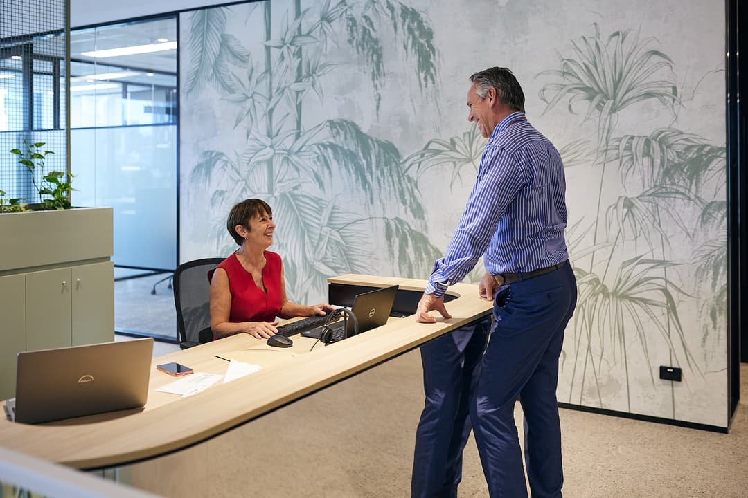 A woman in a red sleeveless top sits at a desk with a laptop, smiling at a man in a blue striped shirt and blue pants who is standing across from her. The room has a nature-themed mural on the wall and modern office furniture.