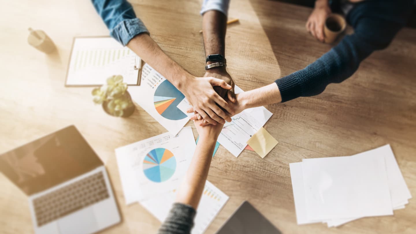 Four people place their hands together over a table scattered with charts, graphs, a laptop, and papers. A small plant is also visible. The scene signifies teamwork and collaboration.