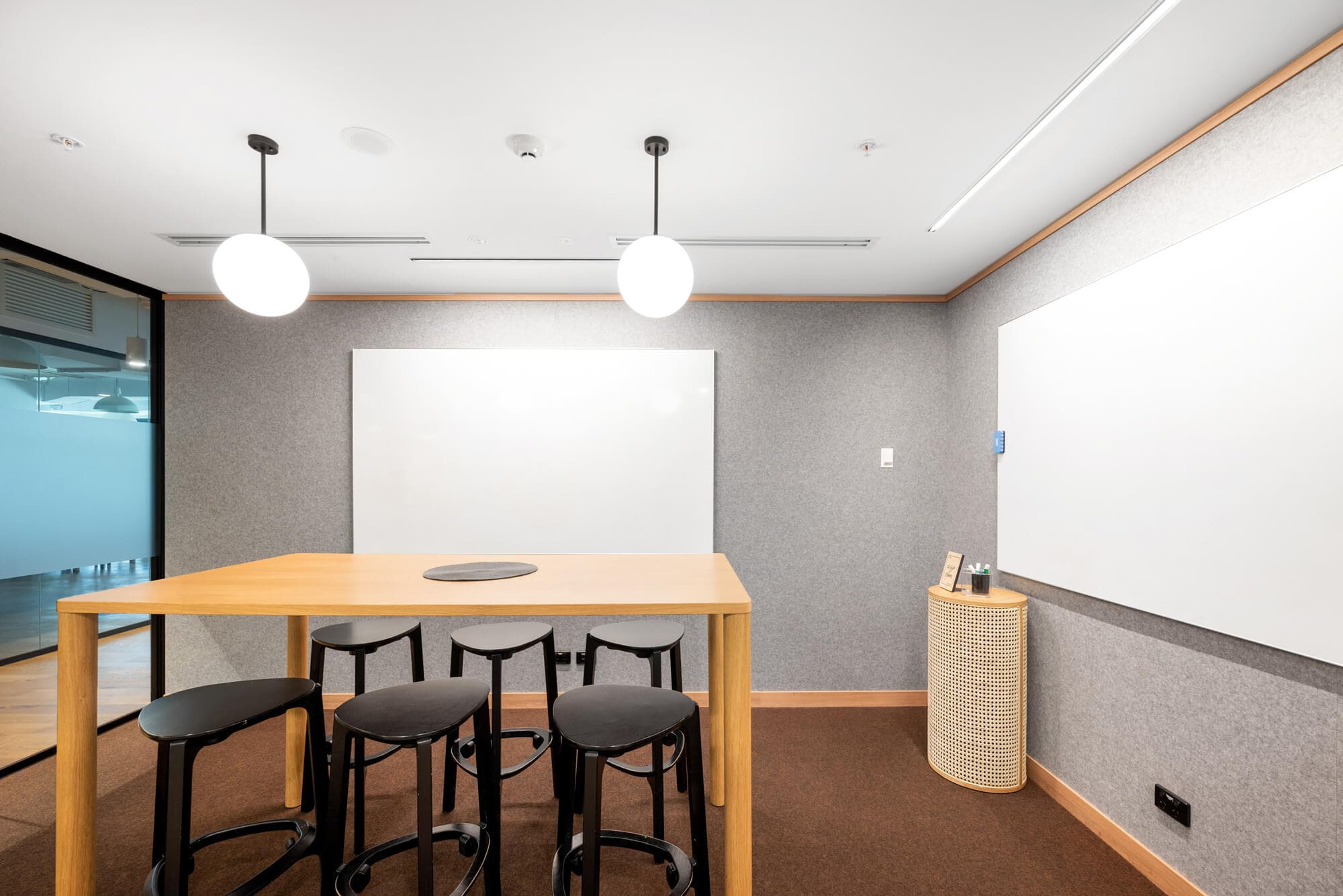 A modern office meeting room with a rectangular wooden table surrounded by four black stools. Two round ceiling lights hang above. A large whiteboard is on the wall, and theres a woven basket with items next to it. The floor is carpeted.