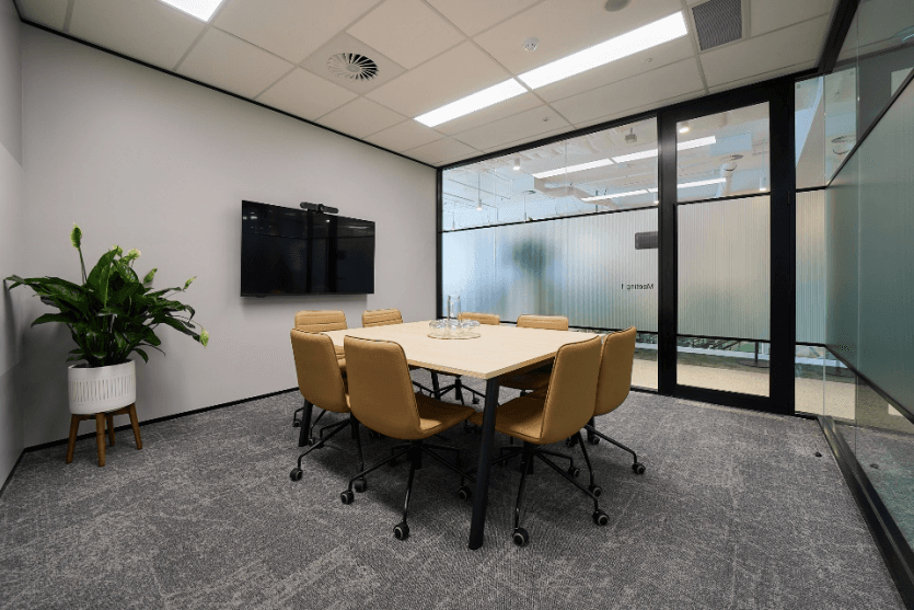 A modern conference room with a rectangular table surrounded by six tan leather chairs. A large flat-screen TV is mounted on the white wall. A potted plant is placed in the corner, and glass walls reveal a hallway outside.