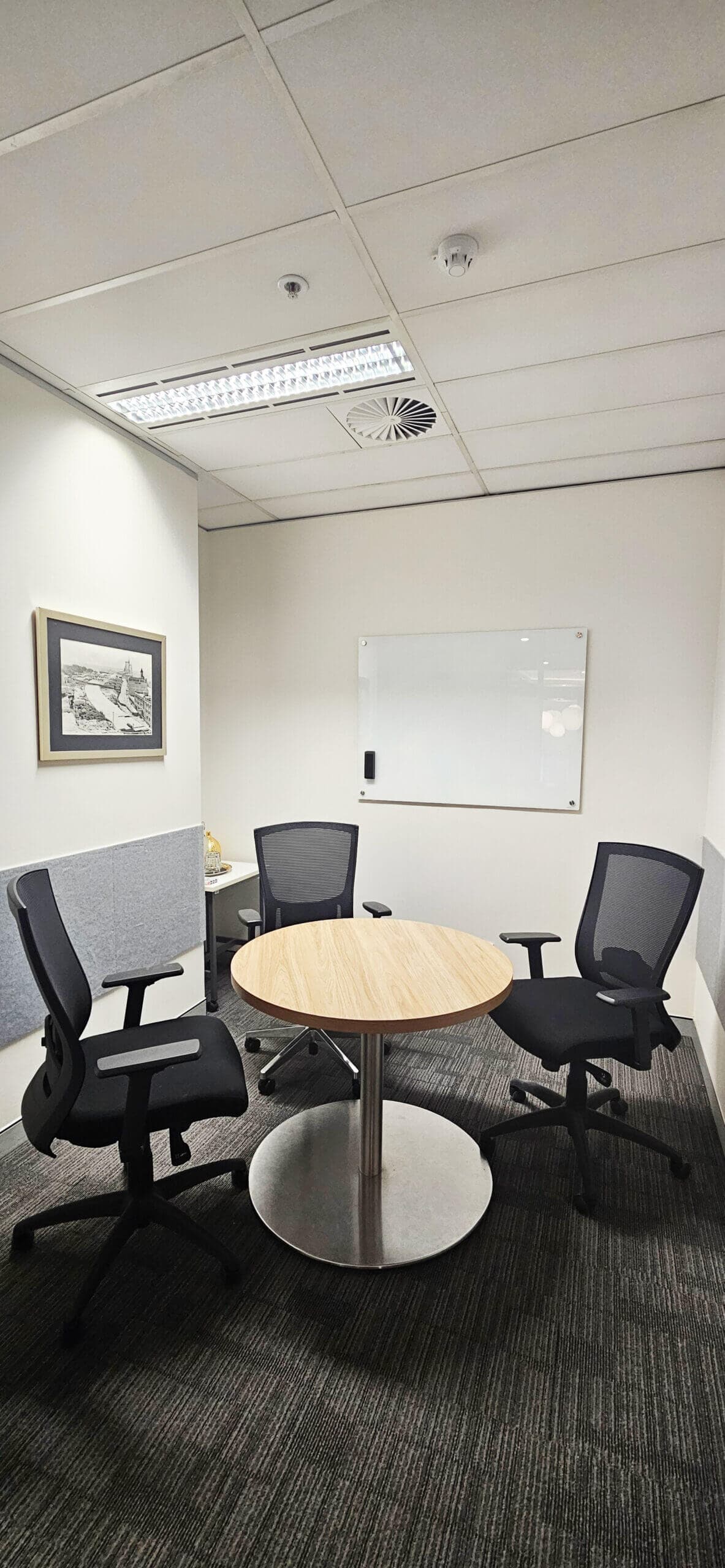 Small office meeting room with a round wooden table, three black mesh chairs, a whiteboard on the wall, and a framed black-and-white picture. The room has neutral colors and fluorescent ceiling lights.