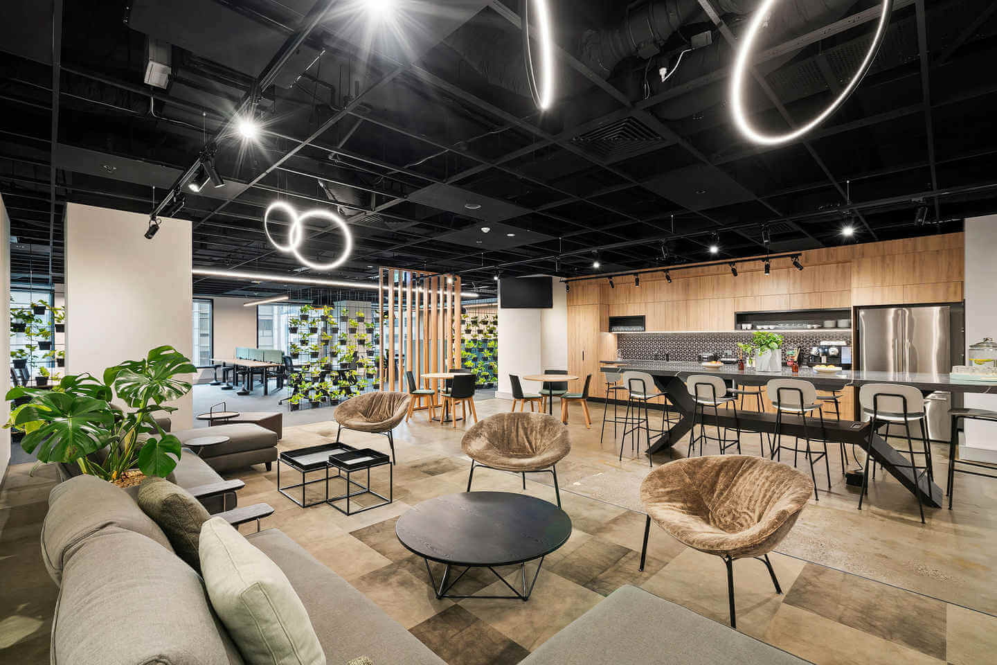 Modern, spacious office space with a lounge area featuring plush chairs, sofas, and round tables. A kitchen with a tiled backsplash and bar seating is in the background. Unique lighting fixtures hang from the ceiling, and a large plant decorates the corner.