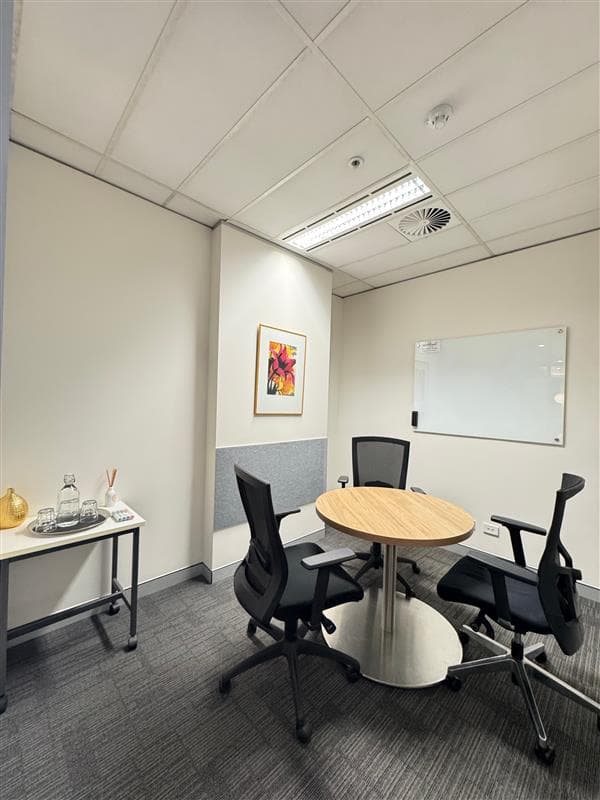 A small office meeting room with a round wooden table, three black chairs, a whiteboard on the wall, a framed flower artwork, and a side table with glasses, a water carafe, and a reed diffuser.
