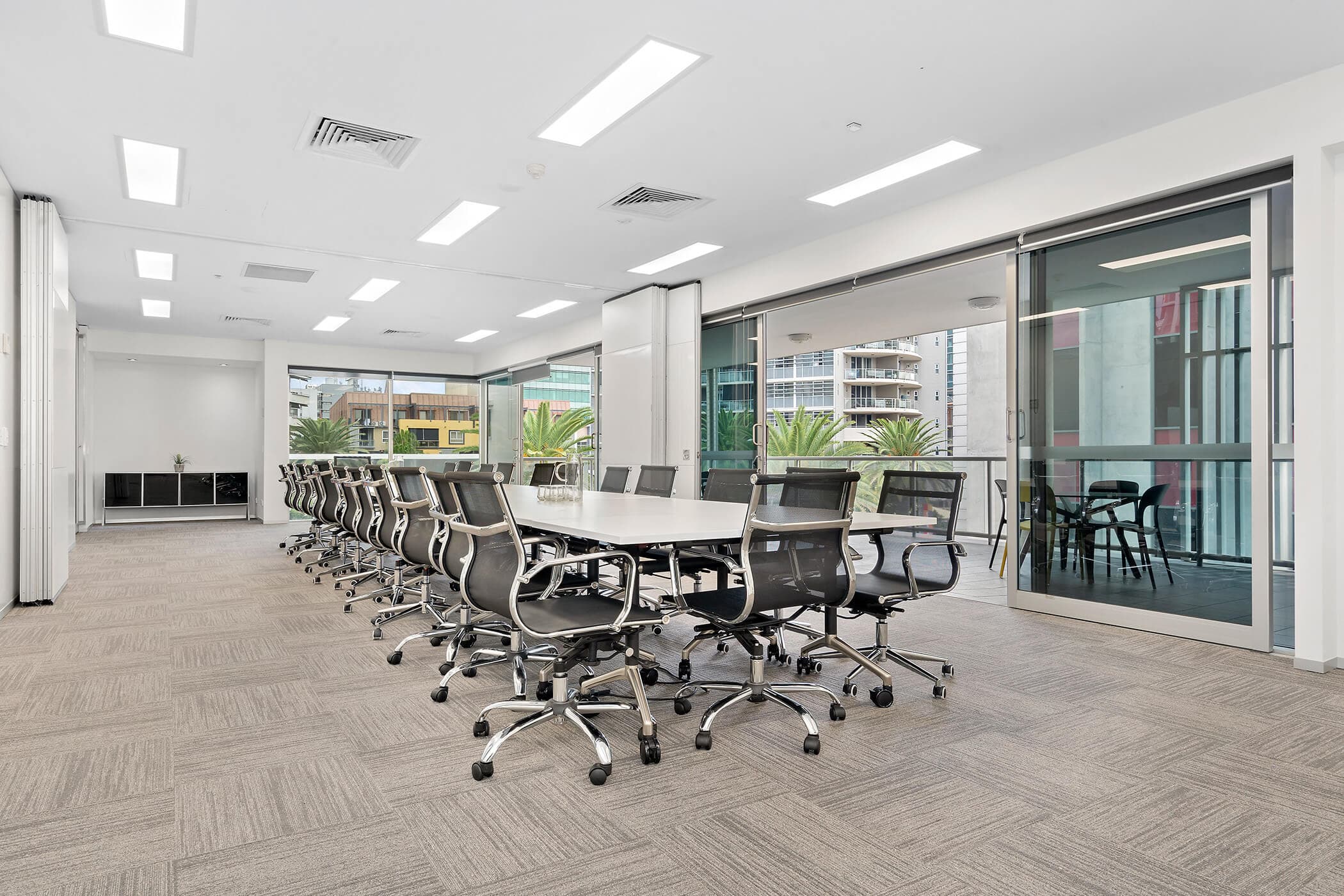 Spacious modern conference room with a long table and multiple swivel chairs. Large windows with sliding glass doors open to a balcony with a view of buildings and palm trees. The room has bright overhead lighting and neutral carpet flooring.