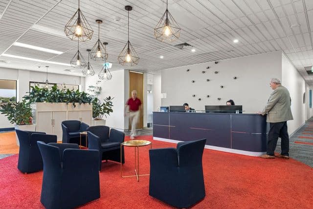 Modern office lobby with navy blue chairs on a red carpet, a small round table, decorative hanging lights, indoor plants, and a reception desk where two people are assisting a standing man. Another person walks toward the lobby.