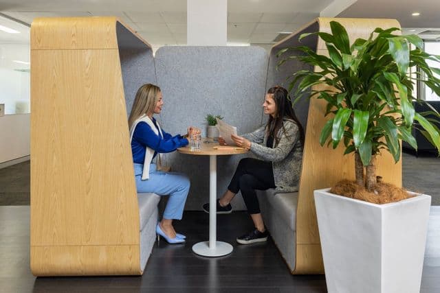 Two women sit across from each other at a round table in a modern office booth, talking and smiling, with a potted plant nearby.