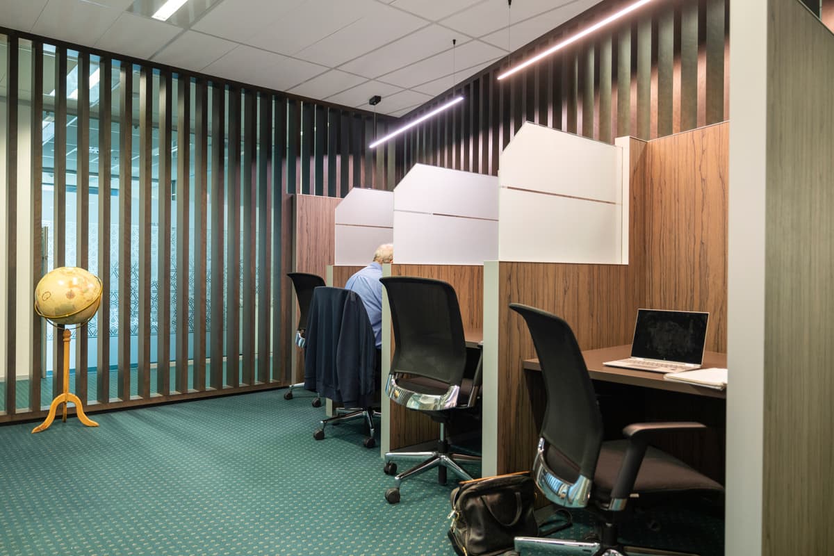 A person sits at a wooden cubicle desk in an office with green carpet, a row of empty desks, a globe on a stand, and a laptop on one of the desks. Vertical glass panels separate the space.
