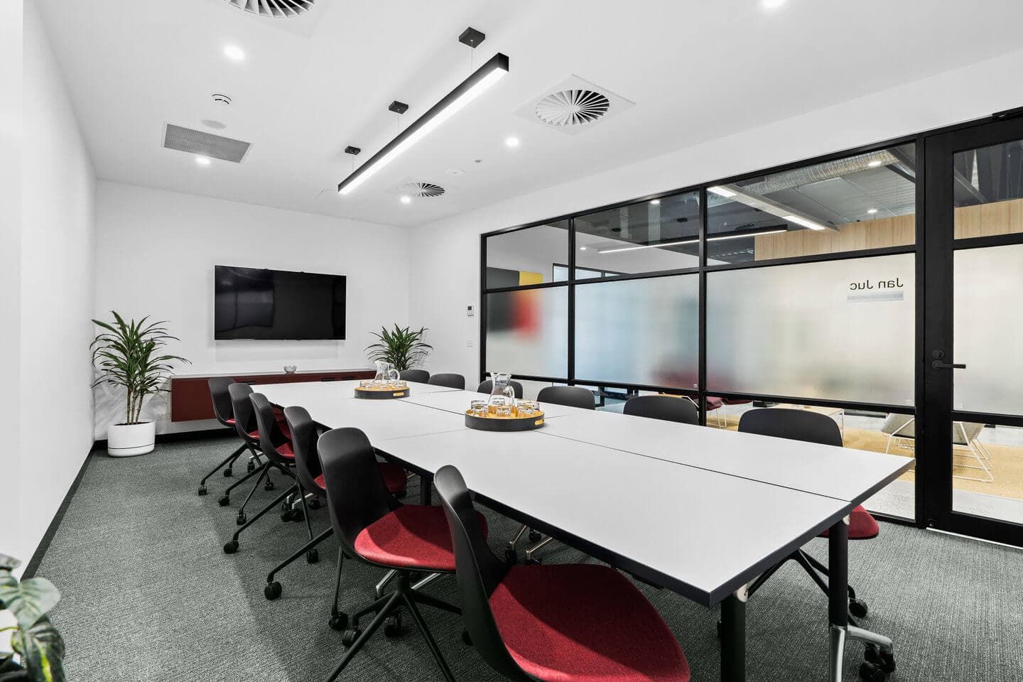 A modern conference room with a long table and red chairs. Two round trays of snacks are on the table. A large TV is mounted on the wall, and there are two potted plants in the corners. Glass doors lead to another room.