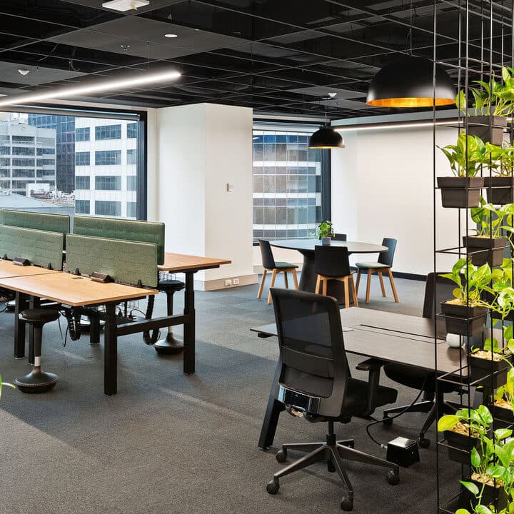 Modern office space with open desks, green dividers, black chairs, round tables, and large windows. Hanging black lamps and potted plants add to the contemporary decor. Cityscape visible through the windows.