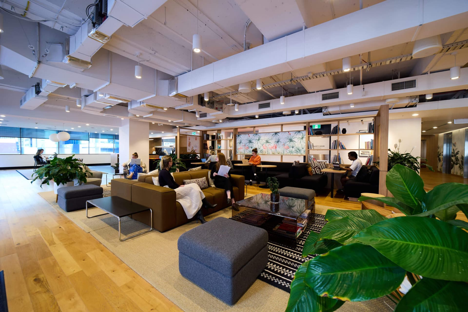 A modern office space with wooden flooring, large green plants, and seating areas featuring sofas and armchairs. People are seated, working or conversing, with bookshelves and decorative art on the walls. Ceiling features exposed beams and ducts.