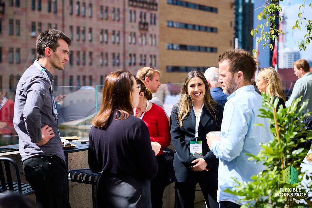 A group of people network and converse outdoors on a sunny rooftop, with city buildings in the background. Everyone appears engaged and smiling. A sign reads Flexible Workspace SF+BA.