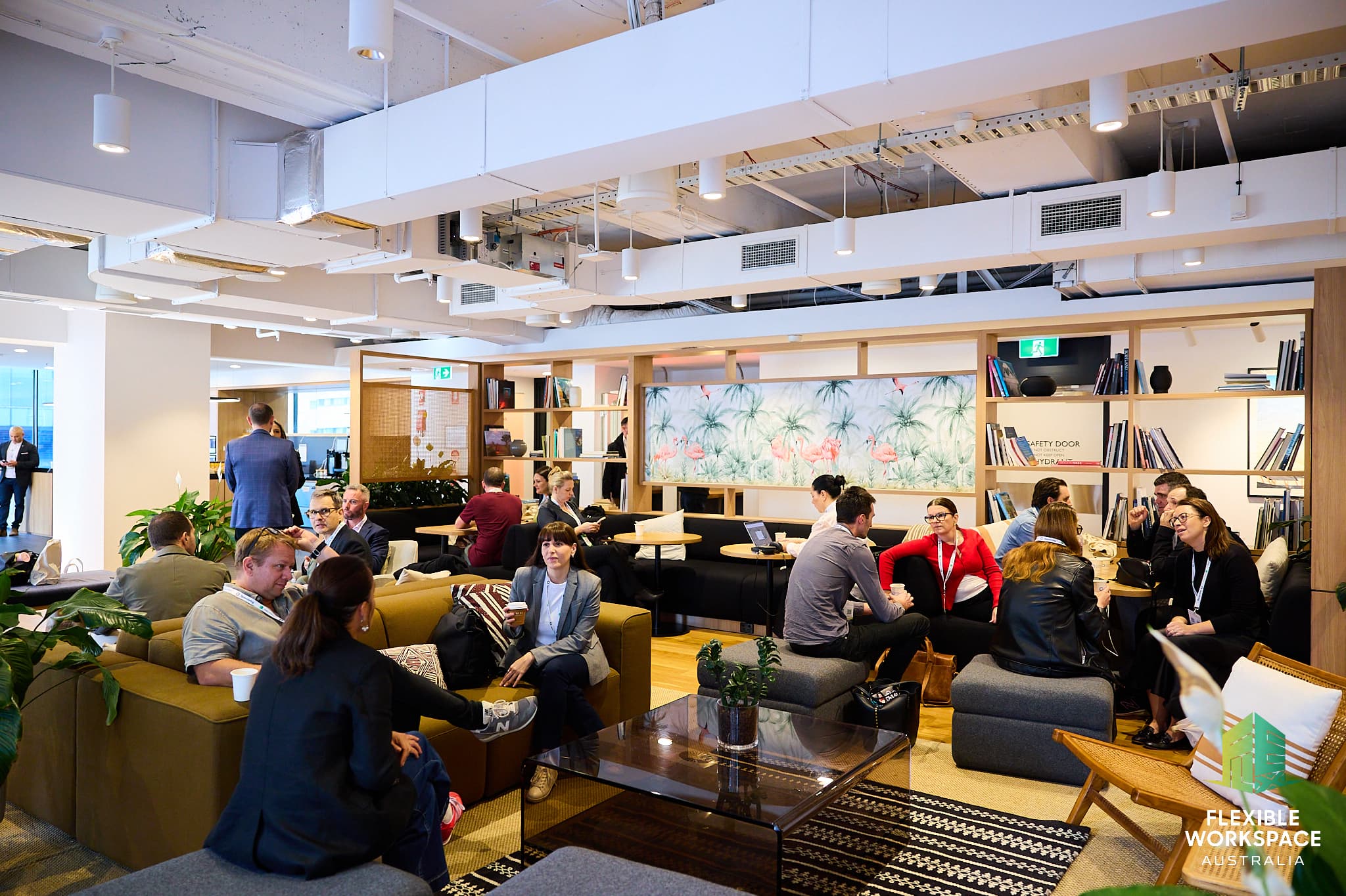 A modern, open-plan office lounge with people sitting on sofas and chairs, engaged in conversations and meetings. Shelves with books, plants, and decorative items line the walls. Bright, welcoming atmosphere.