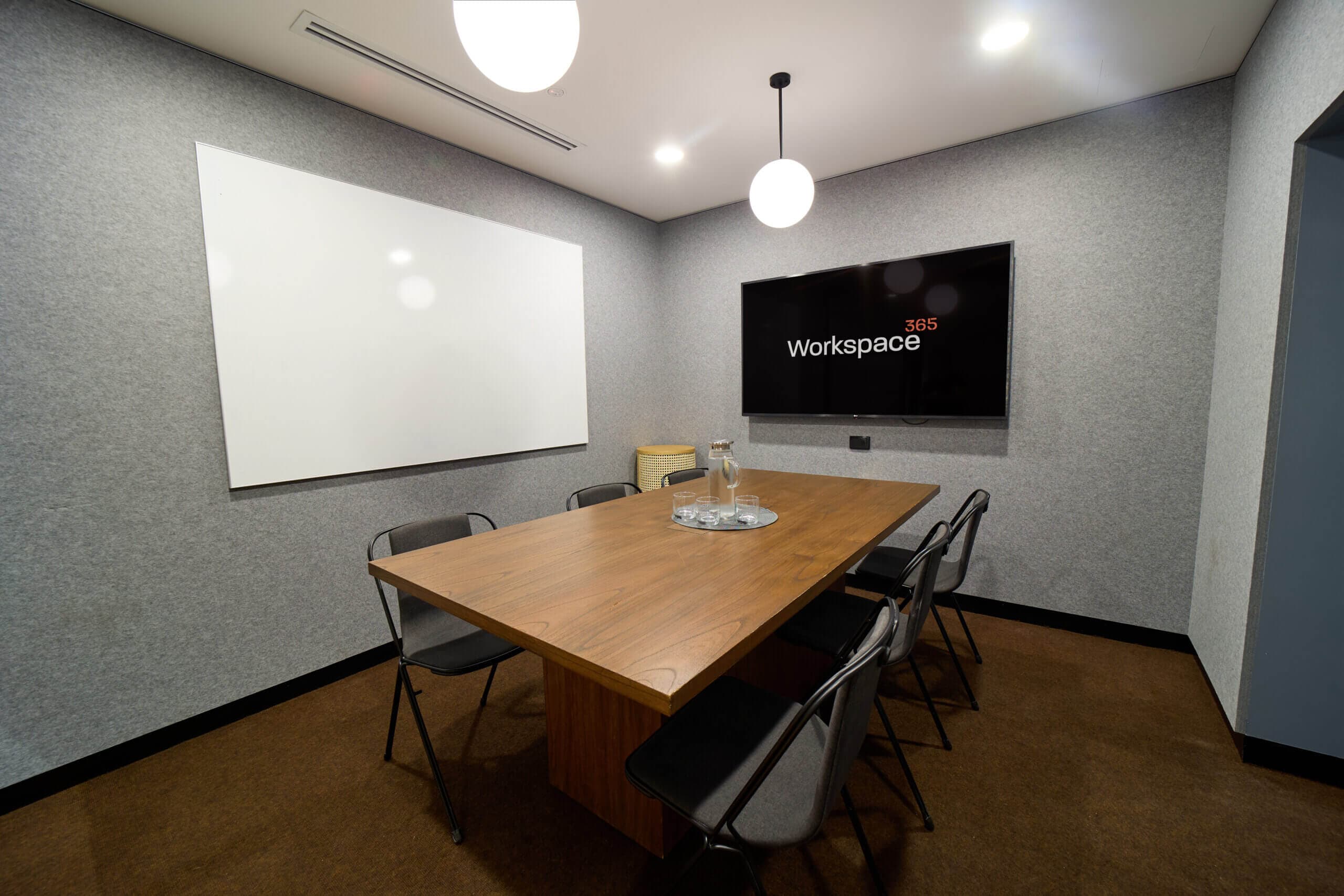 A well-lit meeting room features a wooden conference table surrounded by chairs. A whiteboard and large display are mounted on the wall. A pitcher of water and glasses sit on the table.