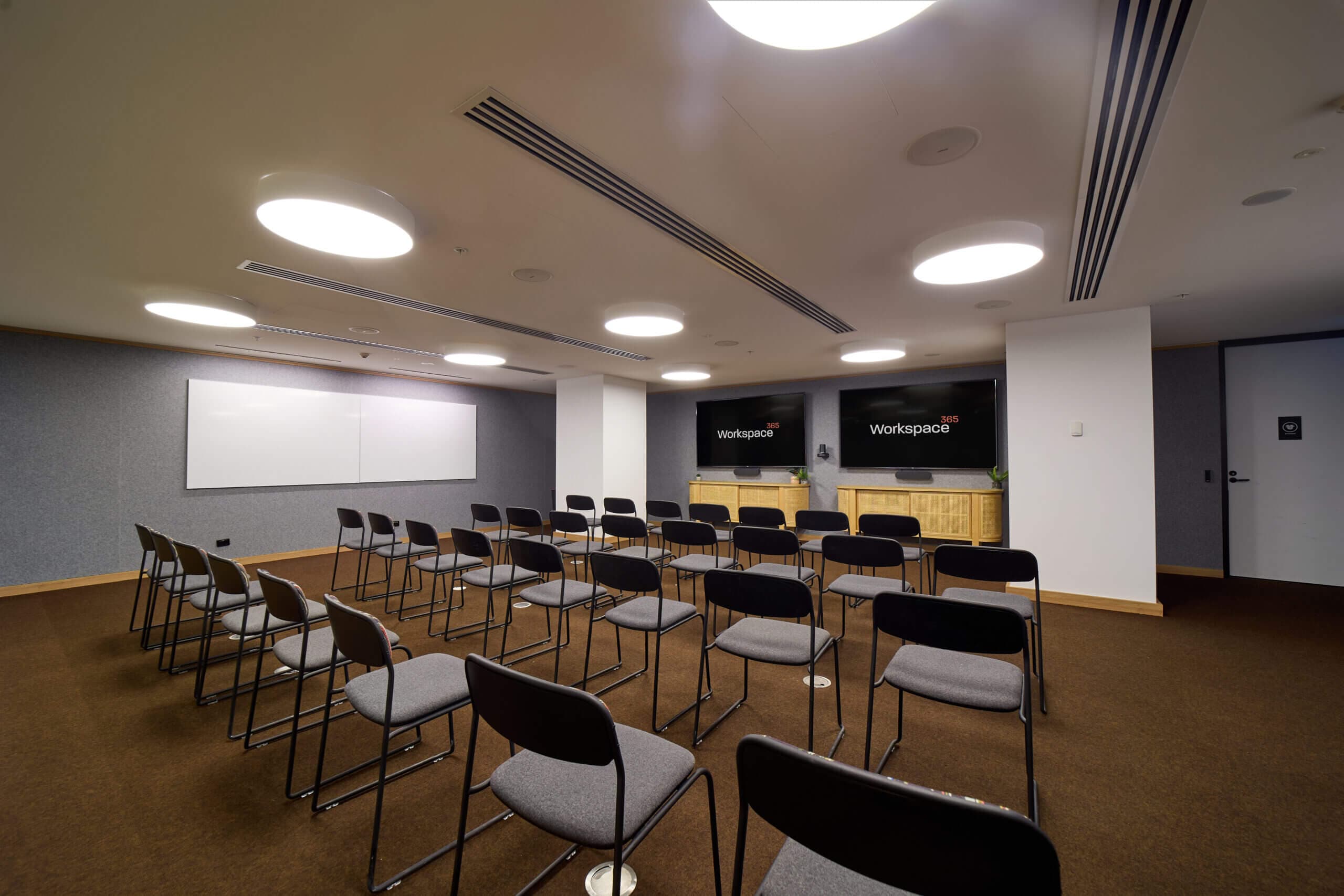 Conference room with rows of chairs facing a whiteboard and two large screens.