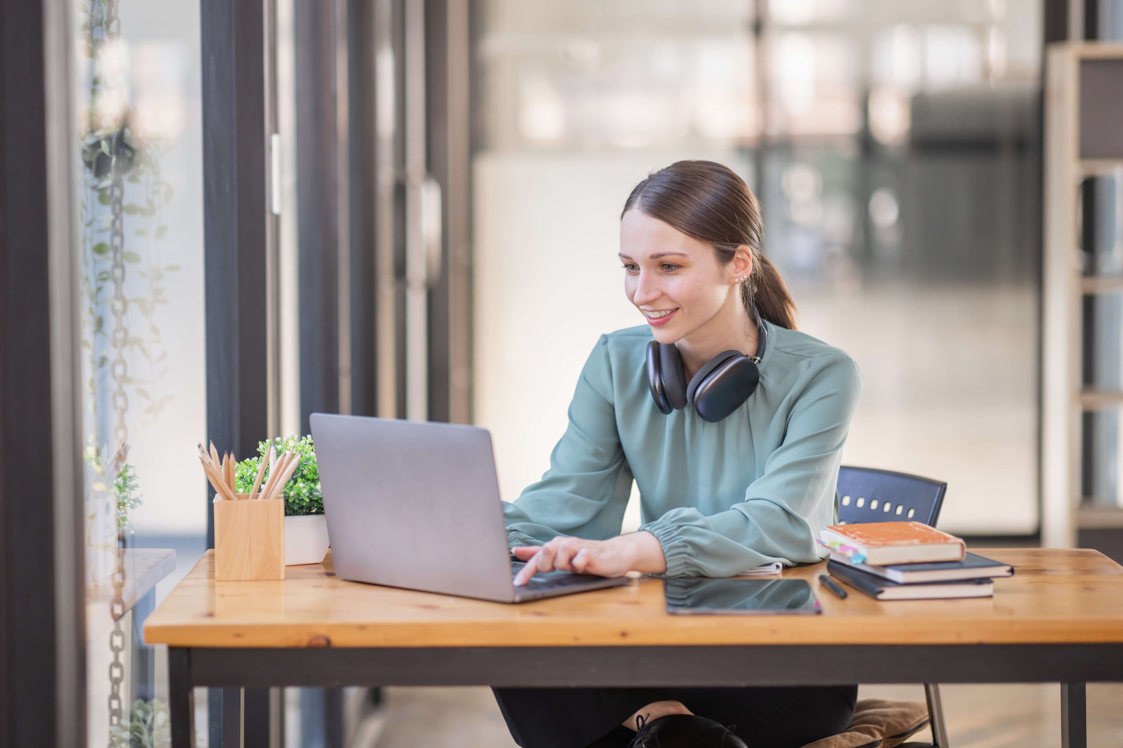 A woman sits at a wooden desk working on a laptop, wearing headphones around her neck. She smiles while focusing on the screen. The desk has books, a tablet, and a potted plant. Large windows in the background let in natural light.