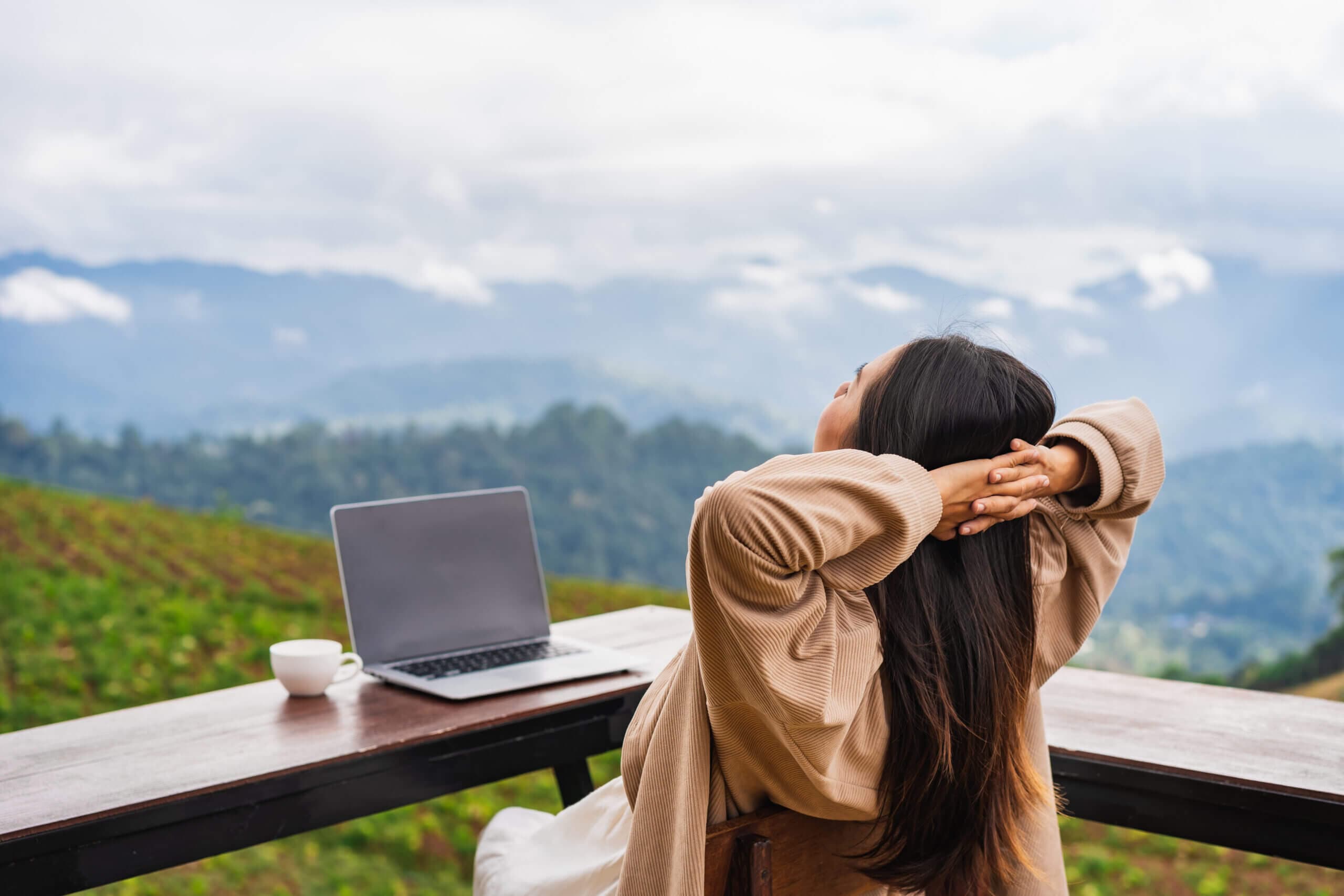 A person with long hair sits at a wooden table on a mountain, stretching with hands behind their head. A laptop and a cup are on the table. The background features cloudy skies and lush green hills.