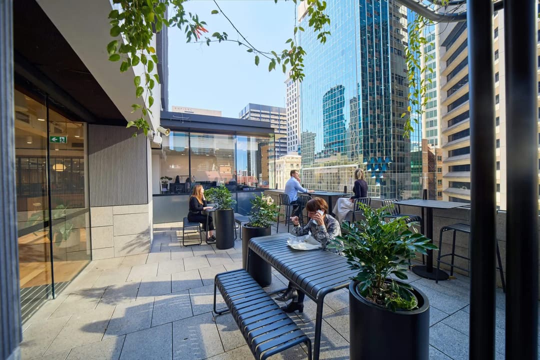 People sit at tables on an outdoor terrace with potted plants and benches, surrounded by tall city buildings with glass facades on a sunny day. Some are working on laptops, while others are talking or relaxing.