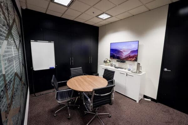 Meeting room with a round table, chairs, whiteboard, and TV displaying a landscape. Modern corporate setting.