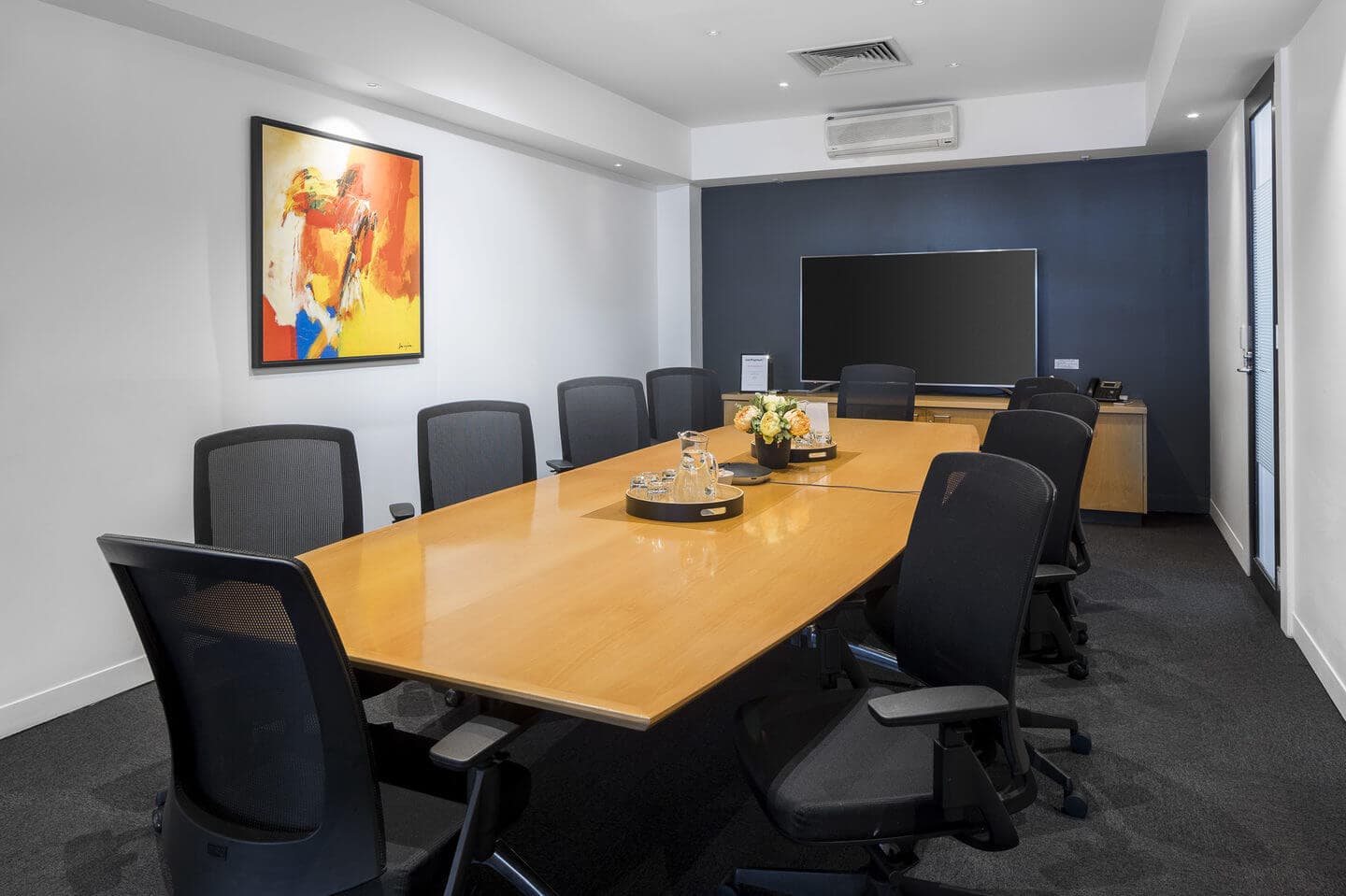 A modern conference room with a long wooden table surrounded by eight black office chairs. A flat-screen TV is mounted on the blue accent wall. A colorful abstract painting hangs on a white wall. A decorative tray with water bottles is on the table.