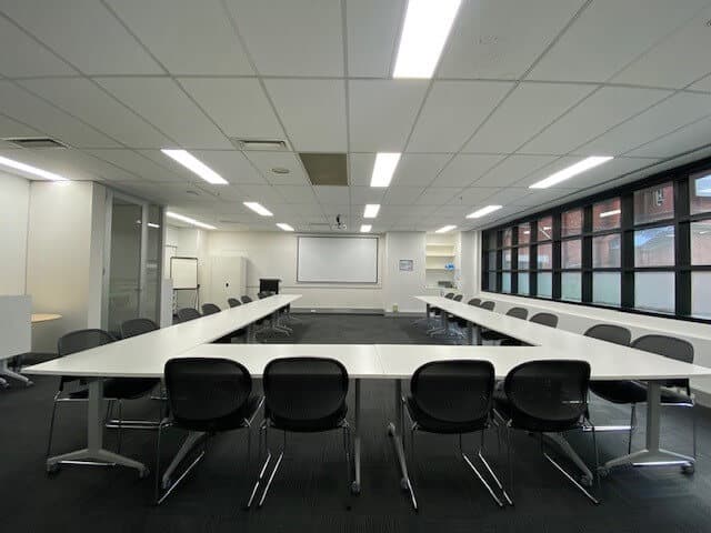 Meeting room with a U-shaped table, black chairs, and a projection screen. Large windows provide natural light.