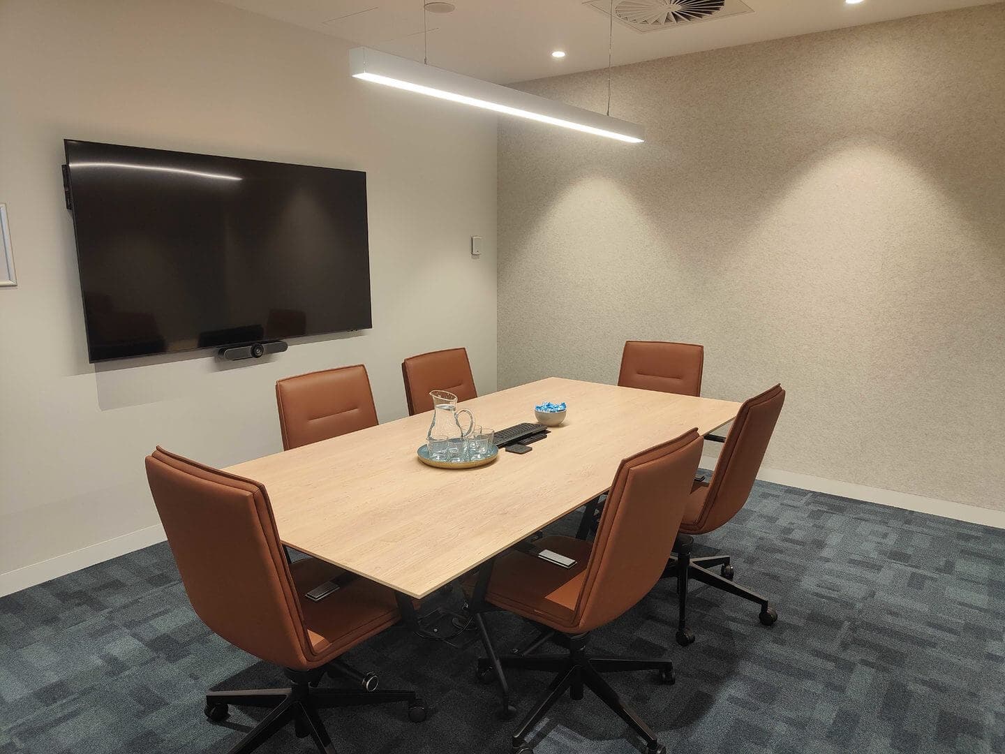 A modern conference room with a rectangular wooden table surrounded by six brown chairs. A flat-screen TV is mounted on the wall. The room has a tray with a glass pitcher and jars, and a minimalist decor with overhead lighting.