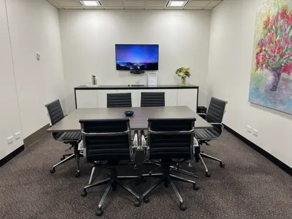 A conference room with a rectangular table and six black chairs. A TV is mounted on the wall displaying a night sky scene. A sideboard with a bouquet of flowers and a colorful painting is visible in the background.