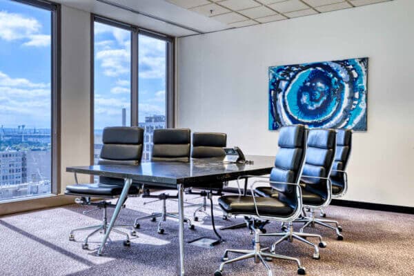 A modern conference room with a long black table and six black leather chairs. Large windows reveal a cityscape and blue sky. A blue abstract painting hangs on the wall, and a phone is placed on the table.
