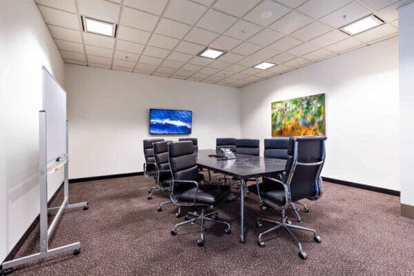 A modern conference room featuring a black table surrounded by eight black office chairs. The room has a whiteboard on one side, a wall-mounted TV showing a landscape image, and a colorful abstract painting on the wall. The carpet is brown.