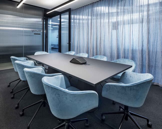Modern conference room with a large black table surrounded by eight light blue upholstered chairs. A speakerphone is placed in the center of the table. The room features a metallic curtain and glass walls, creating a sleek, professional atmosphere.