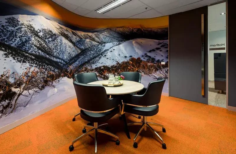 Meeting room with a round table and four chairs on an orange carpet. The wall features a large mural of a snowy mountain landscape. A glass water pitcher and glasses sit on the table. The door is partially open, revealing another room.