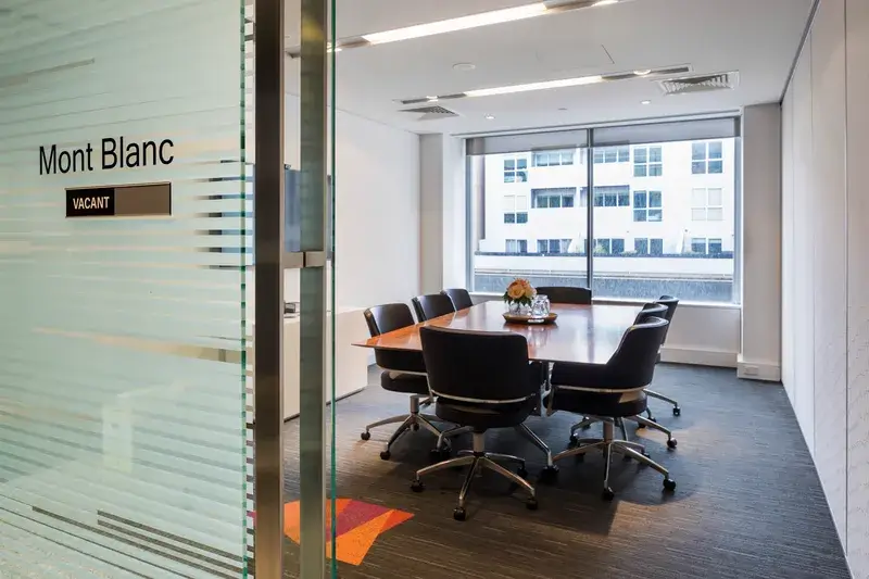 A modern conference room labeled Mont Blanc with a Vacant sign. It features a wooden table surrounded by black chairs, a vase with flowers, large windows, and a glass door.
