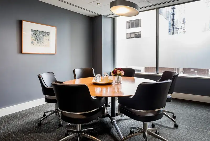 A modern conference room with a round wooden table and five black chairs. A large window shows a view of neighboring buildings. The room has gray walls, a framed artwork, and a ceiling light fixture. A vase with flowers and water glasses are on the table.