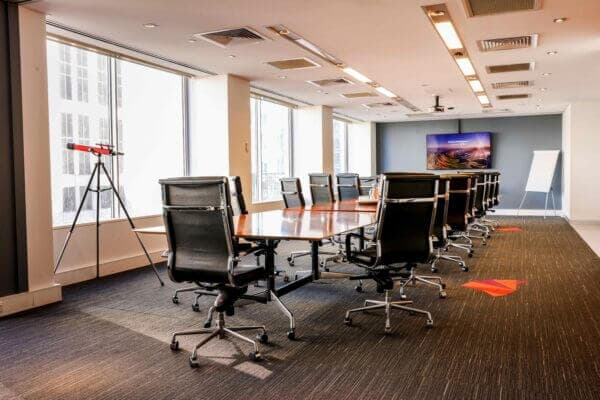 A modern conference room with a long wooden table surrounded by black mesh office chairs. Large windows line the wall, and a tripod with a small telescope stands in the corner. A flat-screen TV and a flip chart are visible at the front.